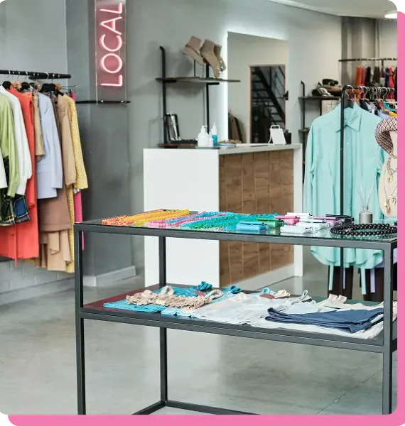 Clothing boutique interior with a two-tier metal display table, colorful garments on racks, and a neon "Local" sign on the wall